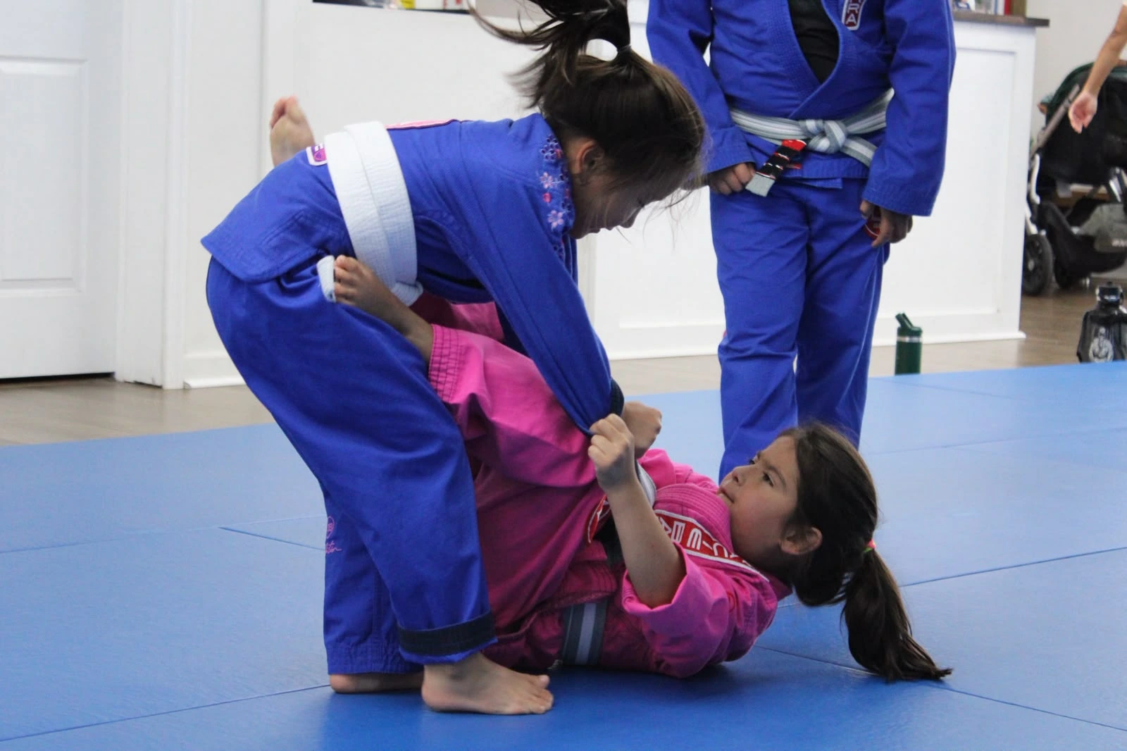 Two girls in blue and pink uniforms practicing Brazilian Jiu-Jitsu together on blue mats